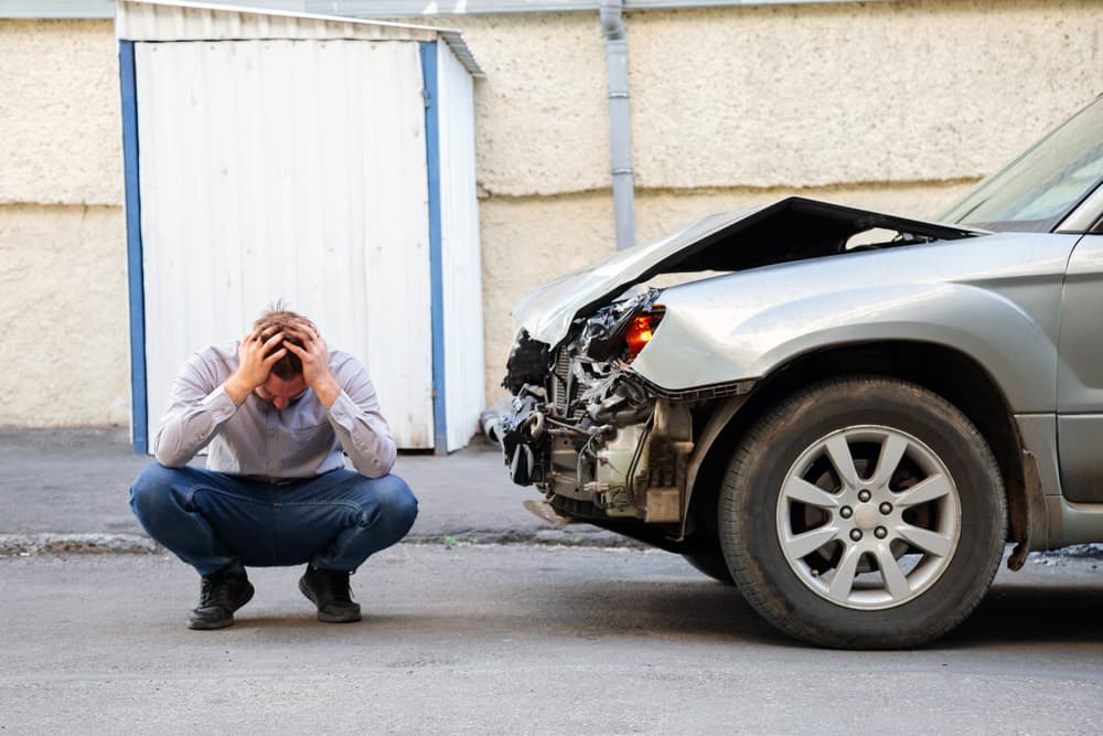 A young man, the driver in a car accident, holding his head near a broken car on the road after the accident.