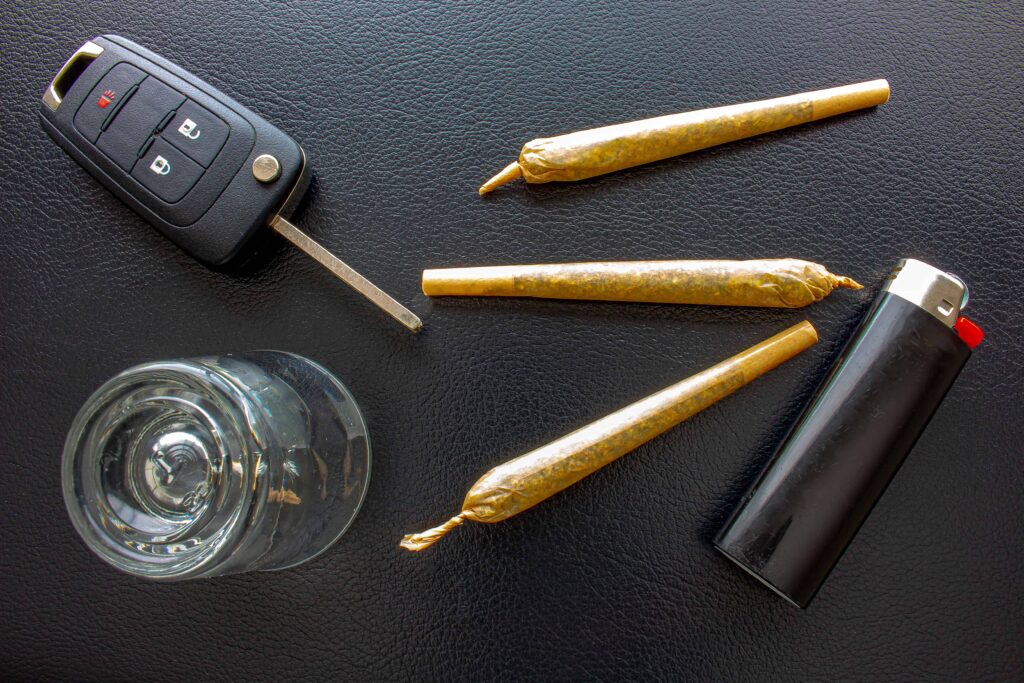 Car key fob beside rolled marijuana joints, a lighter, and a glass on a dark surface, illustrating impaired driving risk