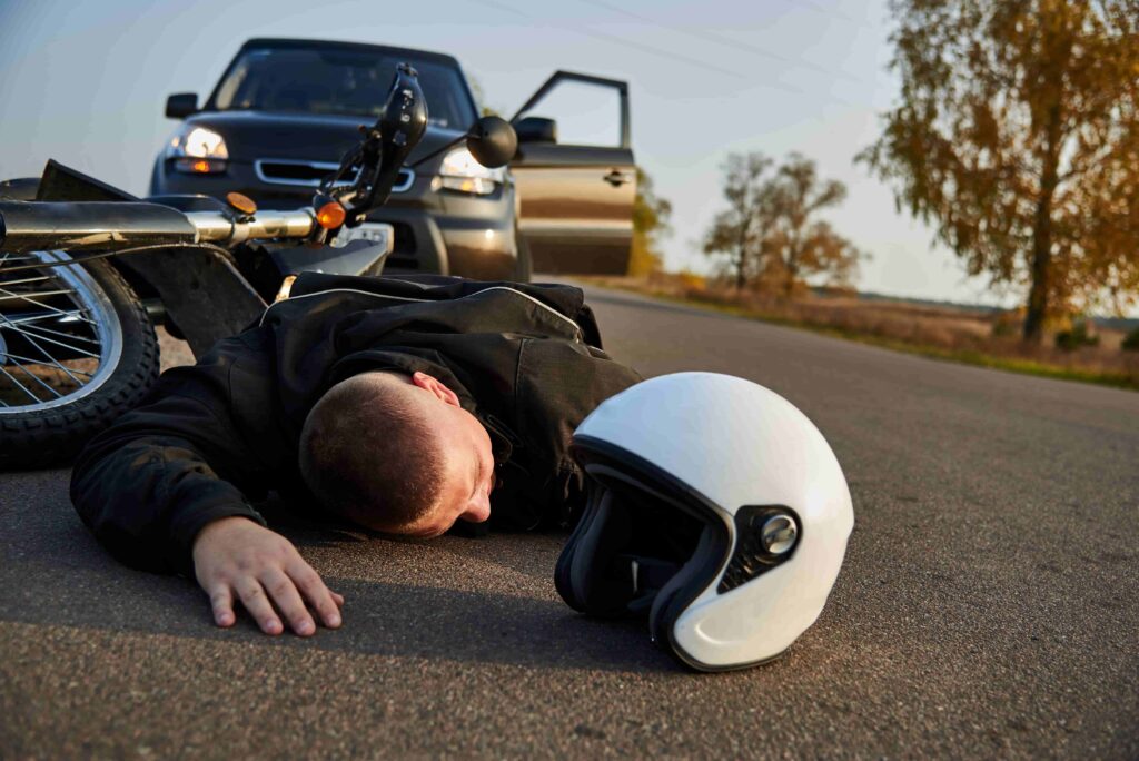 Motorcycle accident victim lying on road after collision with car, helmet and damaged bike nearby.