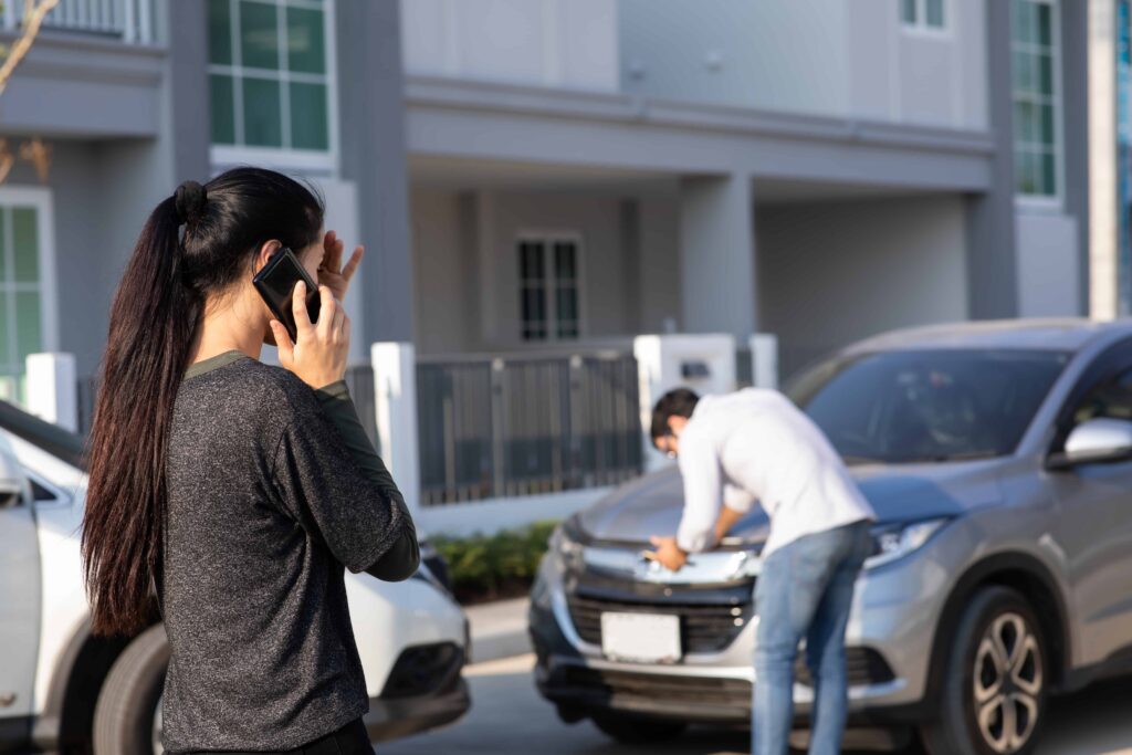 Woman calling for help after a minor car accident while a driver inspects front-end damage on an SUV in a residential street. Woman calling for help after a minor car accident while a driver inspects front-end damage on an SUV in a residential street.