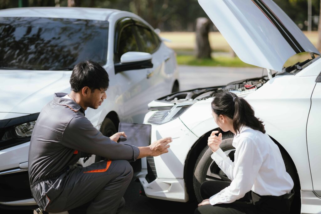 Auto mechanic assessing front-end damage on a white car with the hood open while discussing repair estimate with the driver. Auto mechanic assessing front-end damage on a white car with the hood open while discussing repair estimate with the driver.