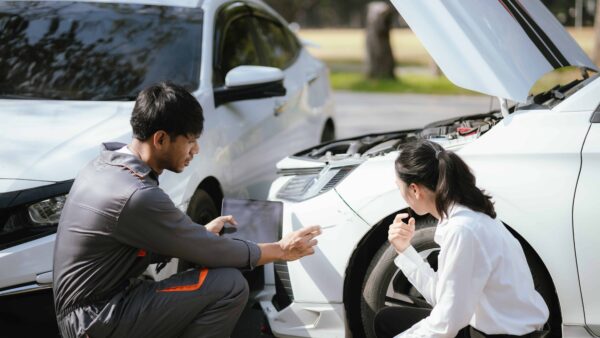 Auto mechanic assessing front-end damage on a white car with the hood open while discussing repair estimate with the driver.