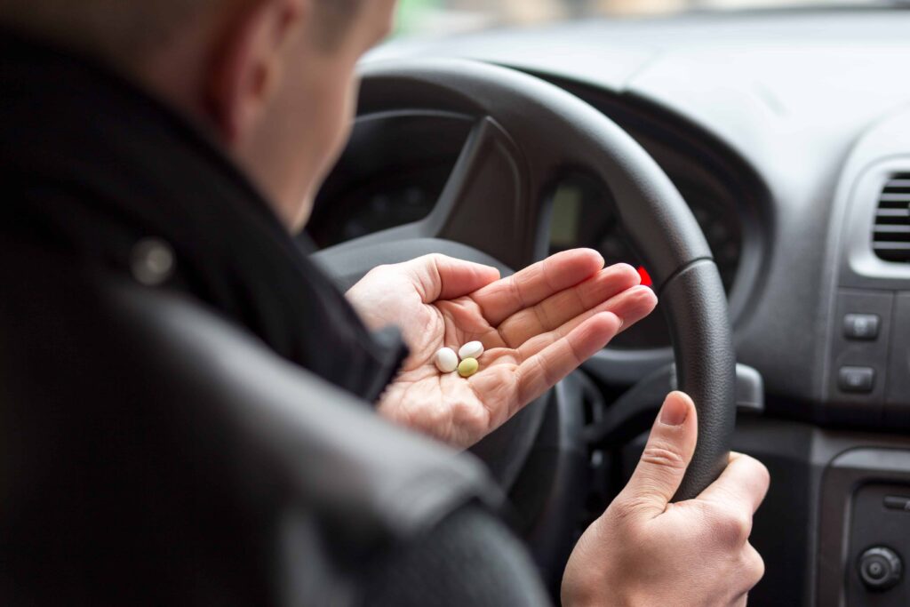 Driver holding pills in hand behind the steering wheel, illustrating the dangers of driving under the influence of medication.